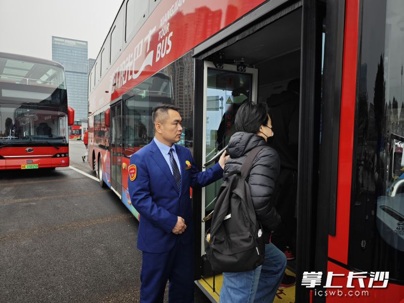 驾驶员匡飞鸽站在等候发车的巴士门前，热情迎接游客。